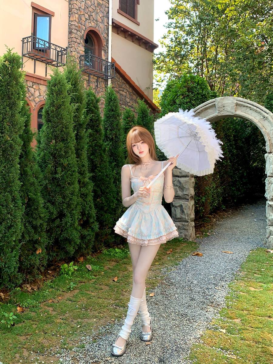 Woman in a light dress holding an umbrella in a garden setting with a stone archway and stone building.