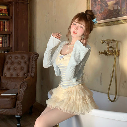 Woman in a white cardigan and beige skirt sitting on a white surface in a room with a bookshelf and painting.