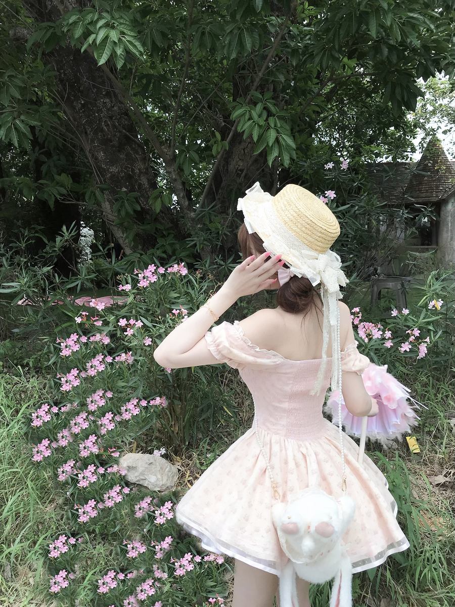 Woman in a pink dress and straw hat standing in a garden with pink flowers and greenery.