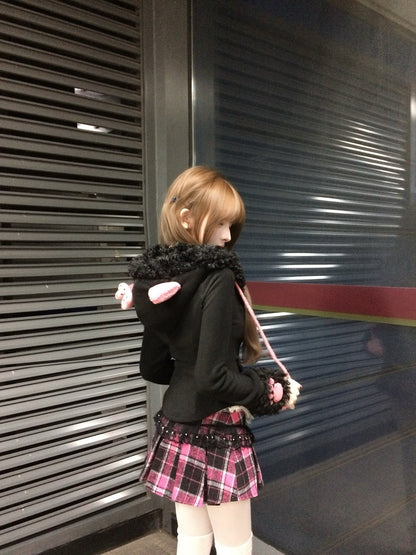 Person wearing a black hoodie with pink accents and a plaid skirt standing in front of a closed shutter.