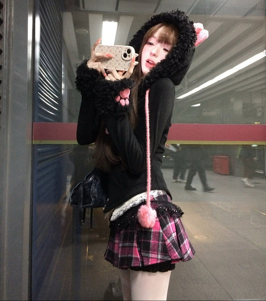 Person taking a mirror selfie in a train station wearing a black coat with a hood and a plaid skirt.