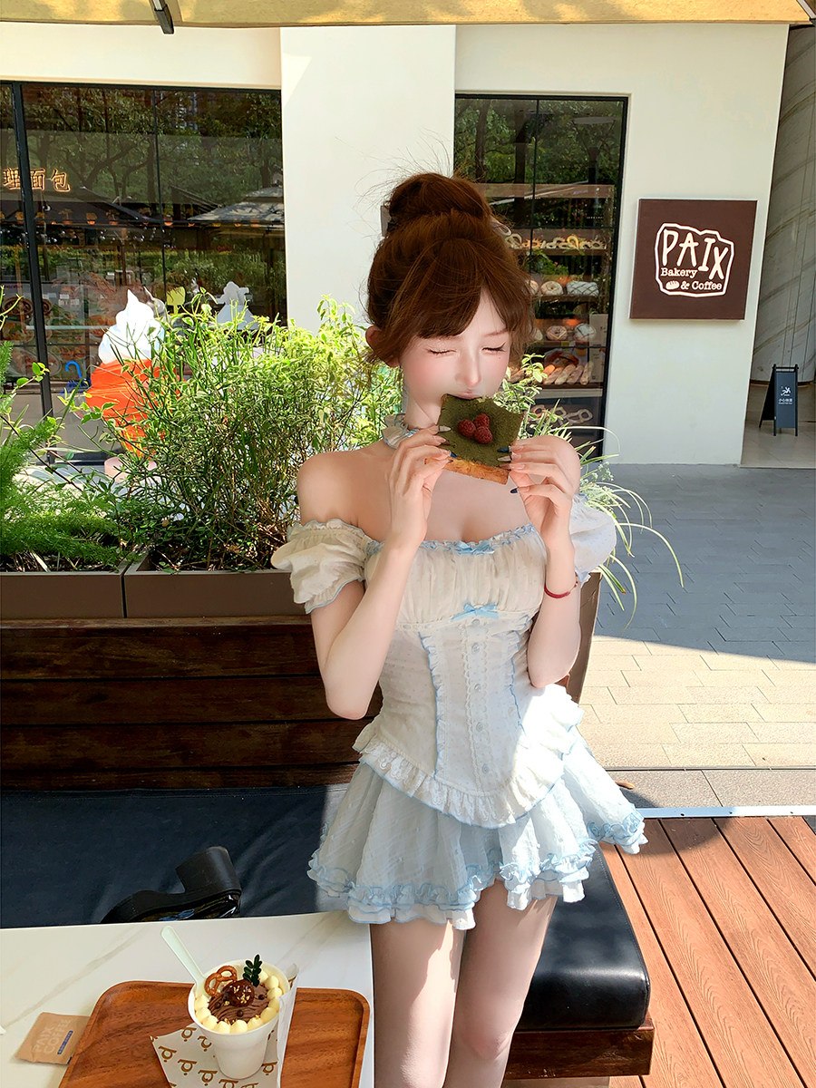 Woman in a light blue dress eating a dessert outdoors with a building and plants in the background.