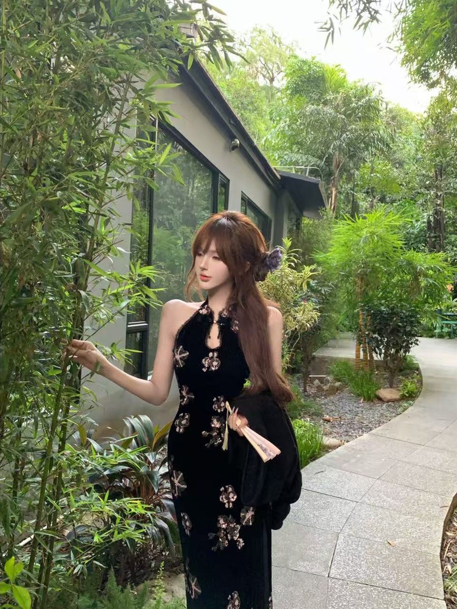 Woman in a black floral dress standing in a garden with greenery and a building in the background