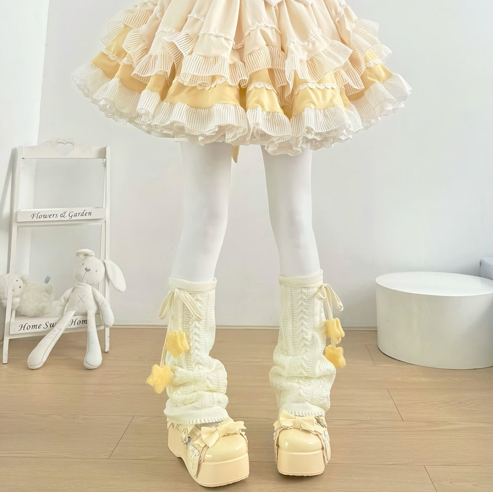 A person wearing a cream and pale yellow ruffled tutu skirt, white tights, and matching cream knit leg warmers with yellow pom-pom accents and chunky platform shoes, standing in a bright indoor room.