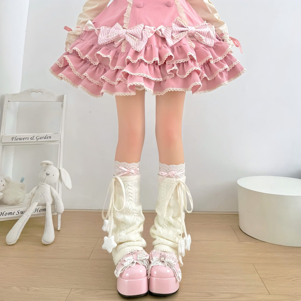 A person wearing a pink ruffled Lolita-style skirt, cream lace-up leg warmers with bows and pom-poms, and pink platform shoes, standing indoors on a wood floor next to a small white chair and stuffed bunny.
