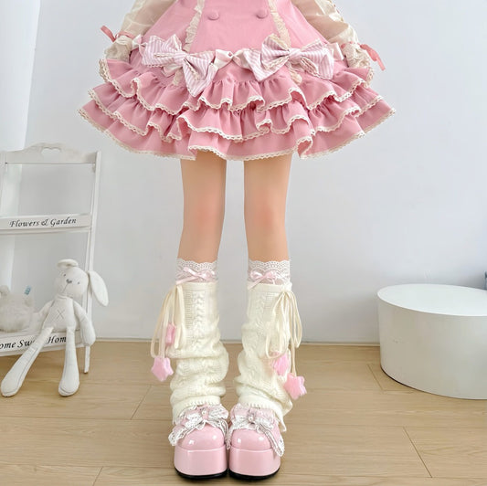 A child's legs wearing a pink ruffled skirt, cream lace-trimmed leg warmers with pink pom-poms, and pink platform shoes, standing in a bright room with a white wall and light wood floor.