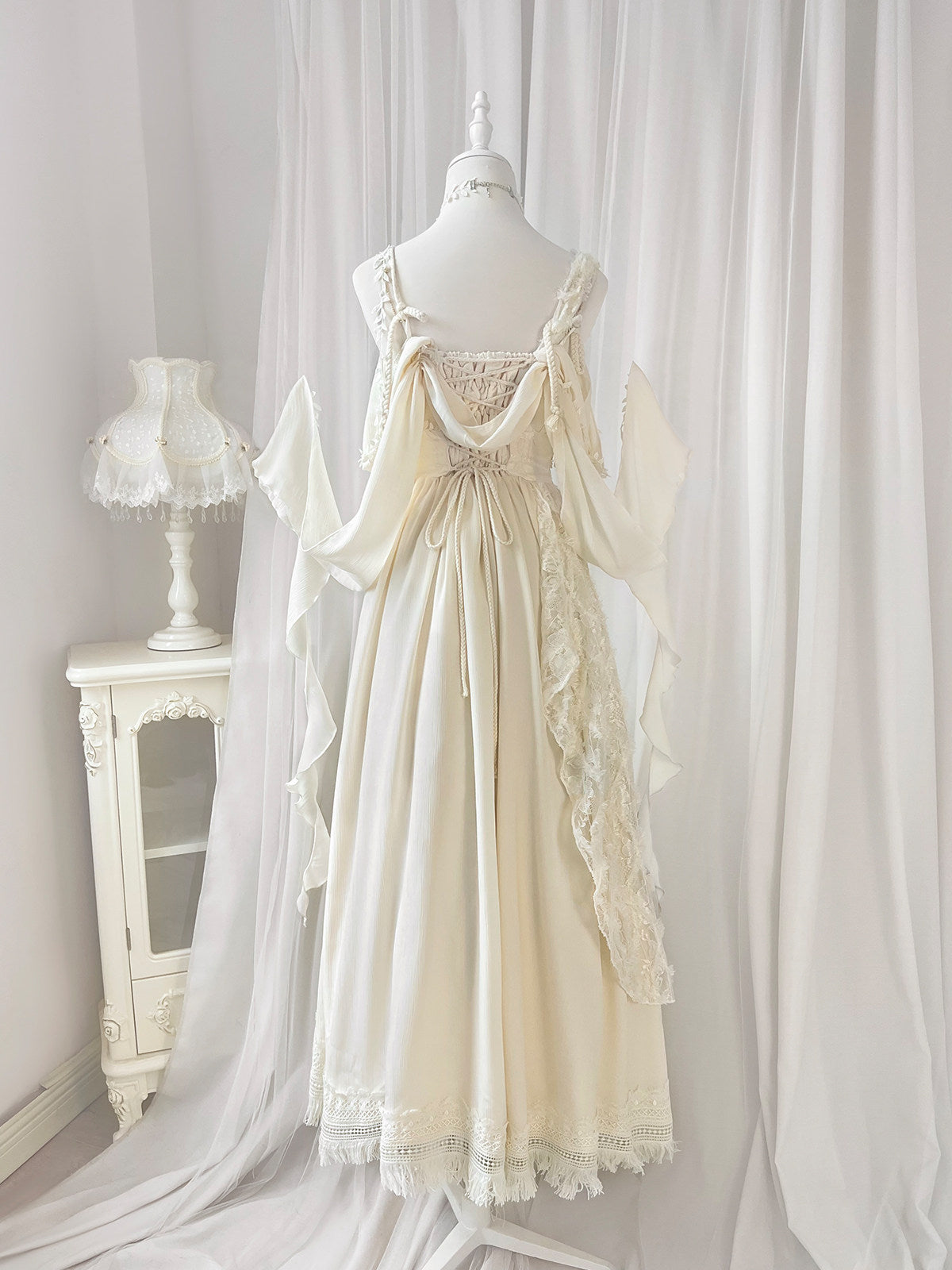 An off-white/cream maxi dress with lace details and ruffled sleeves, displayed on a mannequin in a softly decorated room with white drapes and a small white side table with a lamp.