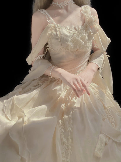 A cream-colored off-the-shoulder ballgown with intricate lace appliques, sheer draped sleeves, and a voluminous ruffled skirt, shown on a model against a solid black background.
