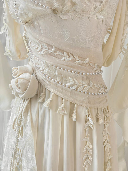 Close-up of a cream-colored embroidered dress featuring intricate floral lace, delicate bead trim, tassels, and a matching fabric rose detail on the skirt.