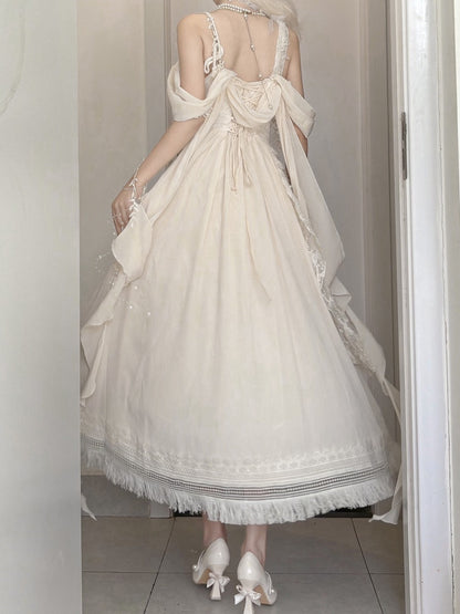 A woman wearing a long cream-colored off-the-shoulder dress with a sheer overlay and lace trim, paired with white heels, standing in a doorway.
