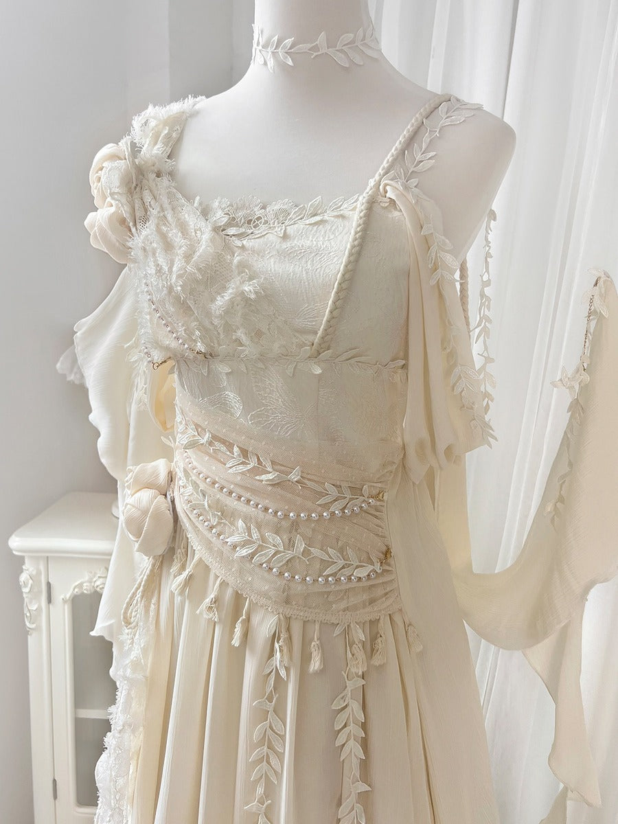 An intricate cream-colored bohemian-style dress displayed on a white mannequin, featuring detailed lace, tassel fringe, floral appliqué, and a pearl-embellished waistband, set against a soft white curtain background.