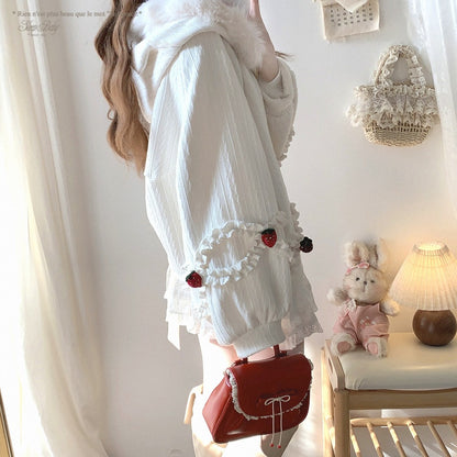 Person wearing a white strawberry jacket with red accents, holding a red bag in a room with a teddy bear and lamp.