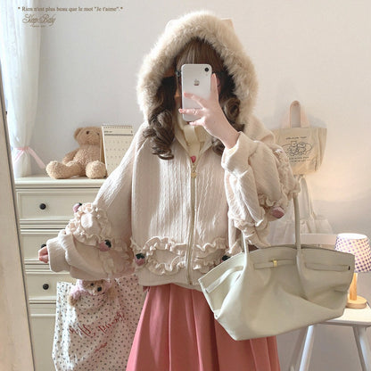 Person wearing a white hooded strawberry jacket with ruffles, holding a beige handbag in a room with a teddy bear and decorative items.