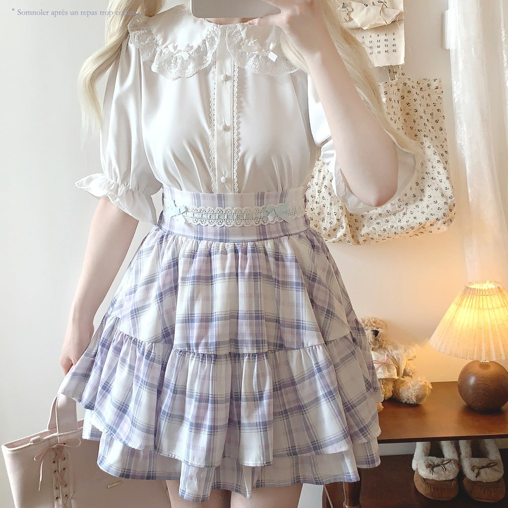 Person wearing a white blouse with ruffled sleeves and a plaid skirt in a room.