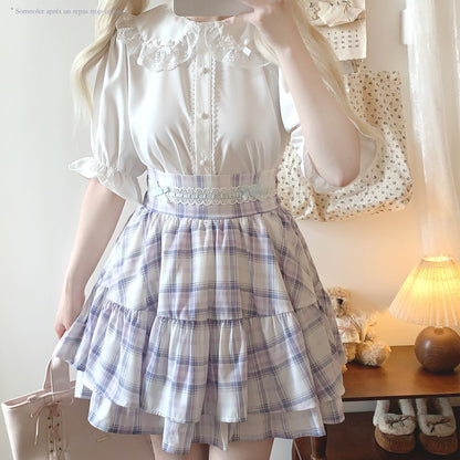 Person wearing a white blouse with ruffled sleeves and a plaid skirt in a room.