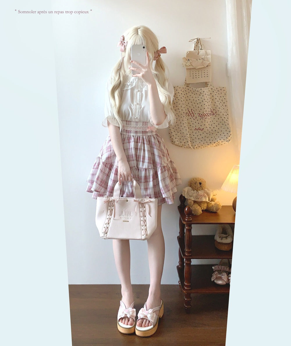 Person wearing a white blouse and plaid skirt with a beige bag in a room.