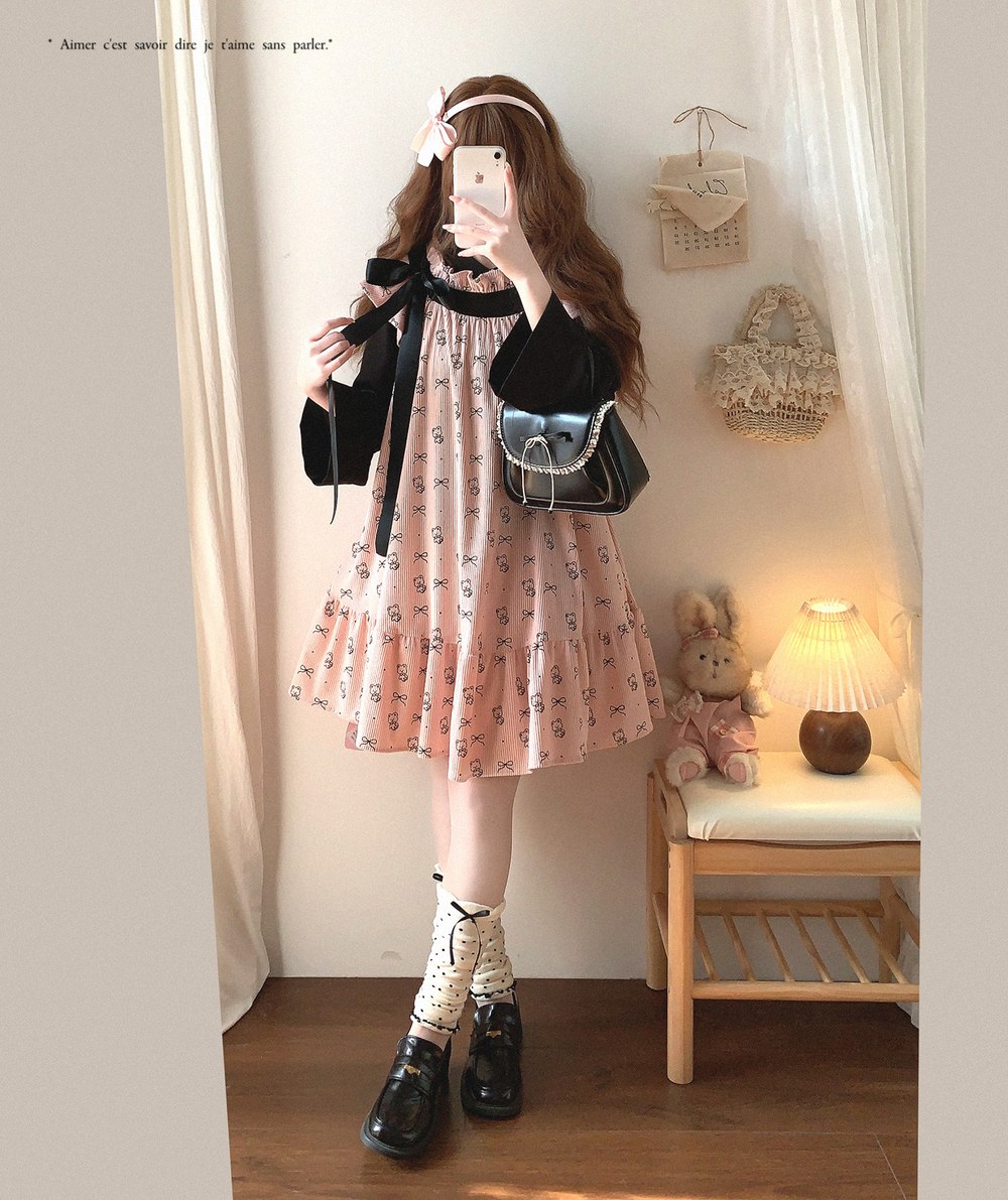 Woman taking a mirror selfie wearing a floral dress and black shoes in a room with a teddy bear and lamp.