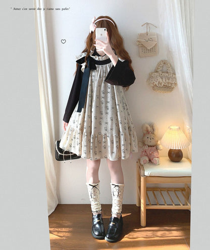 Person wearing a black long-sleeve top and white dress with black text, standing in a room with a lamp and teddy bear.