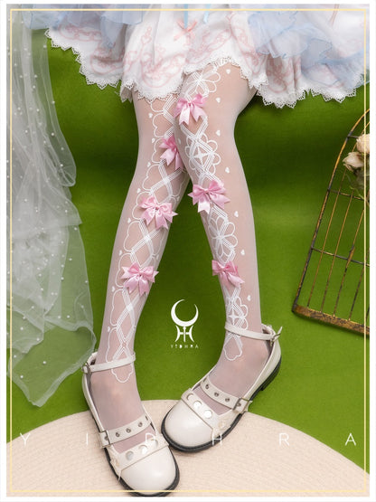 A pair of sheer white tights with pink bow accents and white lace-like patterns, worn by a model in a white frilly skirt and white Mary Jane-style shoes, set against a bright green background.