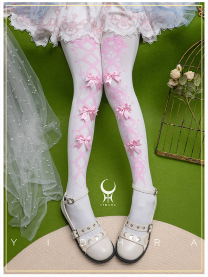 White and pink patterned Lolita-style tights with pink bows, worn by a model with a white ruffled skirt and white lace-up shoes, set against a green background.