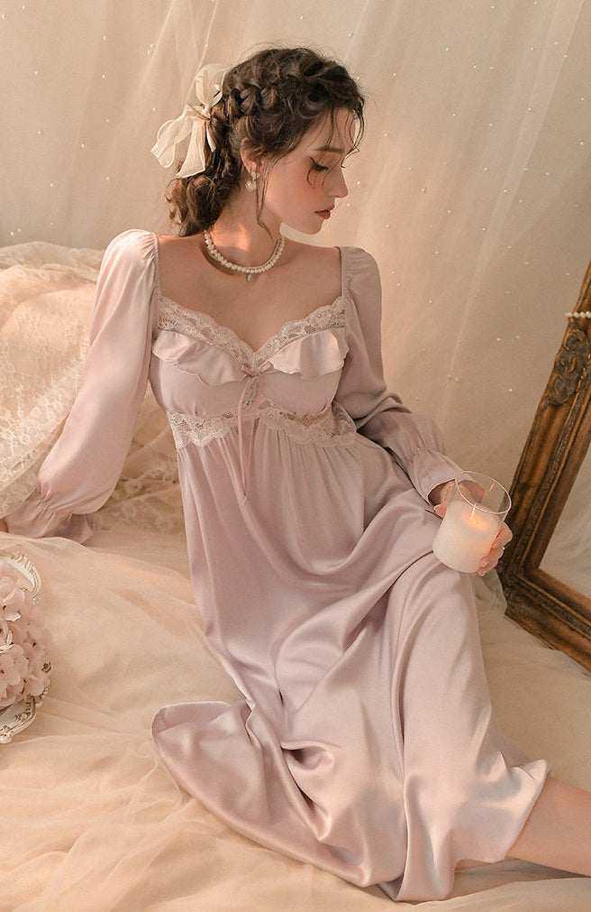 A woman wearing a long, light pink satin nightgown with lace trim sits on a bed in a softly lit bedroom setting, holding a lit candle.