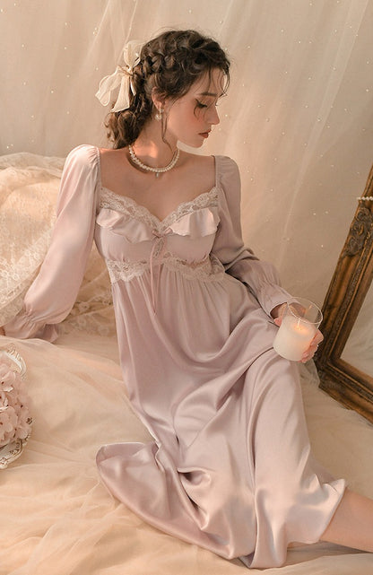 A woman wearing a long, light pink satin nightgown with lace trim sits on a bed in a softly lit bedroom setting, holding a lit candle.