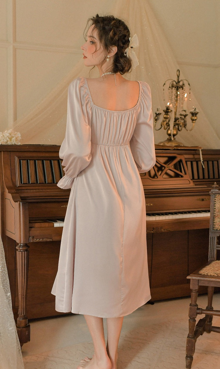 A woman stands with her back to the camera wearing a pale blush pink midi dress with long puff sleeves and a smocked bodice, posed in front of a vintage wooden piano in a softly lit room.