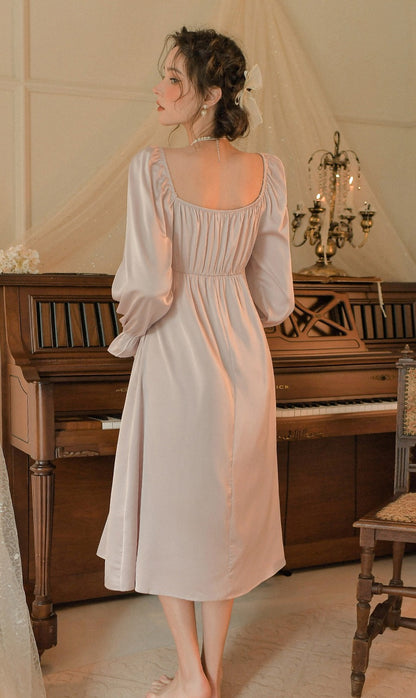 A woman stands with her back to the camera wearing a pale blush pink midi dress with long puff sleeves and a smocked bodice, posed in front of a vintage wooden piano in a softly lit room.
