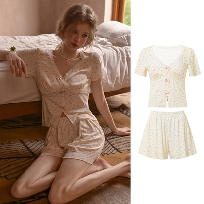 A two-piece short-sleeve pajama set shown on a model kneeling on a bed and also as a clean product composite on the right. The pajama set features a light cream base with a tiny pink floral print and small pink bow details on the chest.