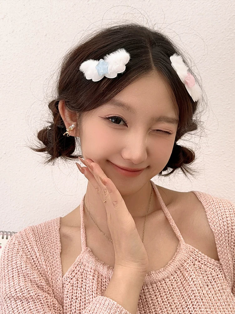 A woman with her hair styled in two half-up buns wearing fluffy white hair clips, a pink knit top, and small gold hoop earrings, posing against a textured off-white wall.