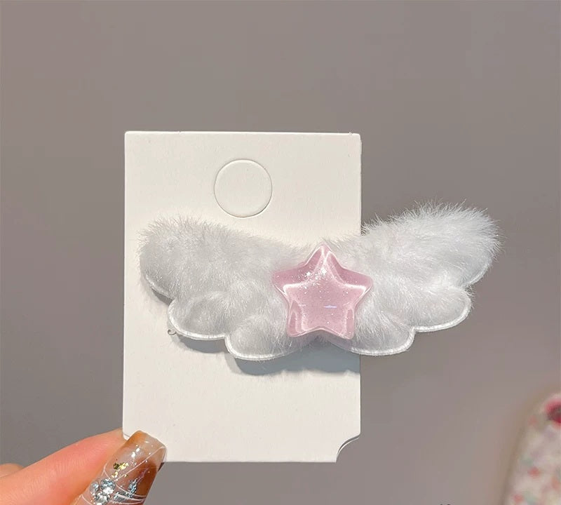 A white fluffy angel wing hair clip with a translucent pink star in the center, attached to a white display card and held by a hand with glittery nails.