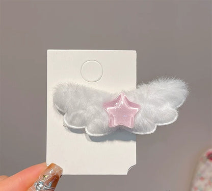 A white fluffy angel wing hair clip with a translucent pink star in the center, attached to a white display card and held by a hand with glittery nails.