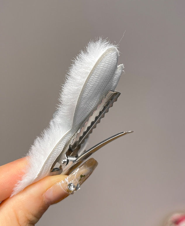 A close-up of a hand holding a white faux fur hair claw clip with a silver metal spring clip mechanism against a soft gray background.