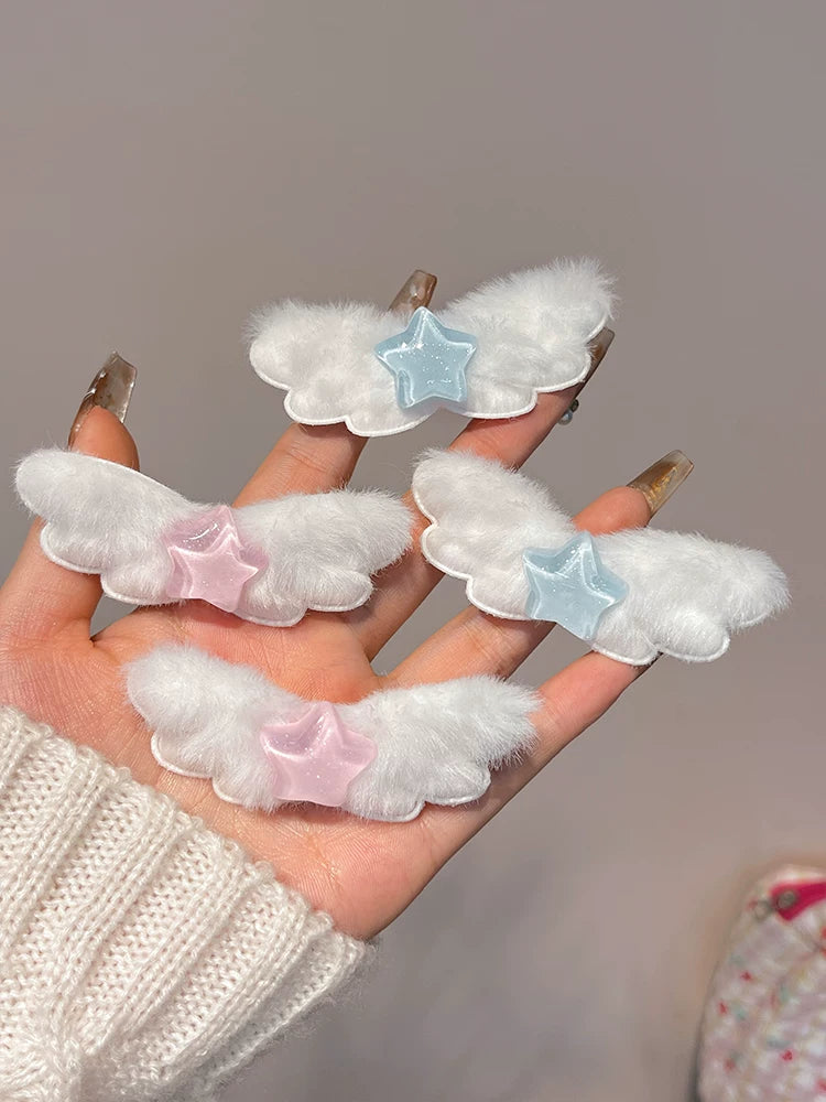 Four white fluffy angel wing hair clips with pastel pink and light blue star accents, held in a hand wearing a cream knit sweater.
