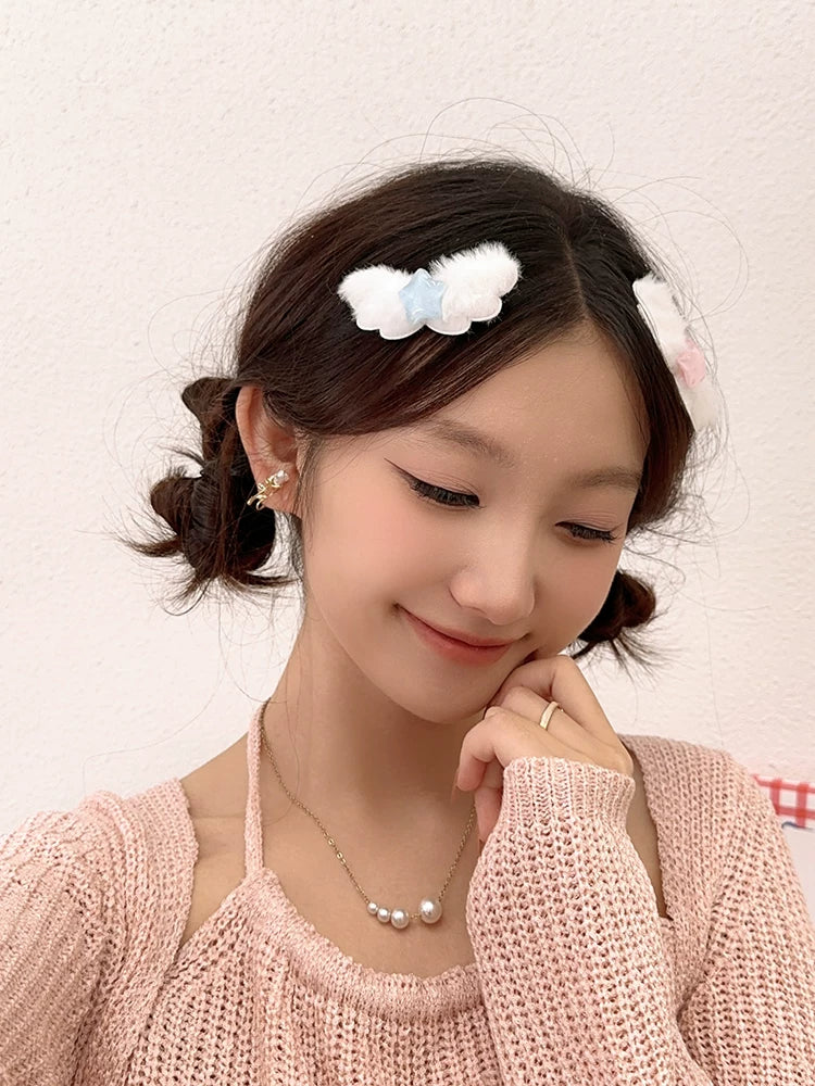 A young woman with her hair styled in half-buns wears fluffy white and light blue bow hair clips on the sides of her head. She is wearing a pink knit sweater and a pearl necklace against a plain, light-colored wall.