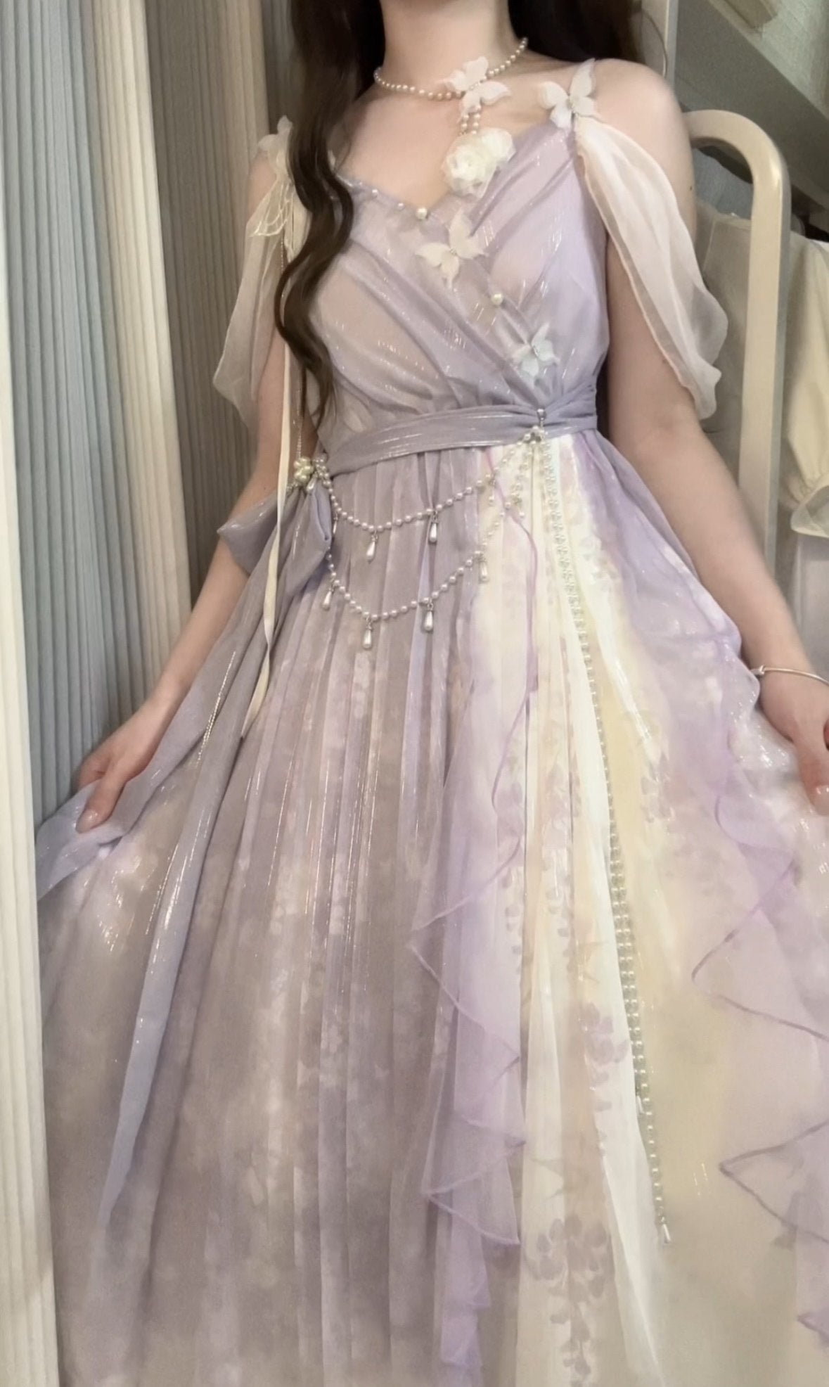 A woman wearing a long, flowing lavender and cream tulle dress with floral embroidery, sheer flutter sleeves, and a beaded waist chain, photographed indoors.