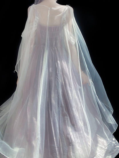 A sheer white tulle cape or drape worn over a long, light lavender dress, shown from the back against a solid black background.