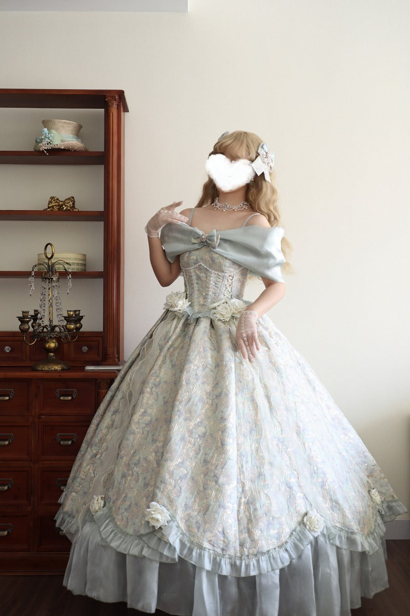A woman wearing an elaborate off-the-shoulder ball gown with a light blue-gray ruffled overlay and a pale floral lace skirt, standing indoors in front of a cream wall and a dark wood cabinet.