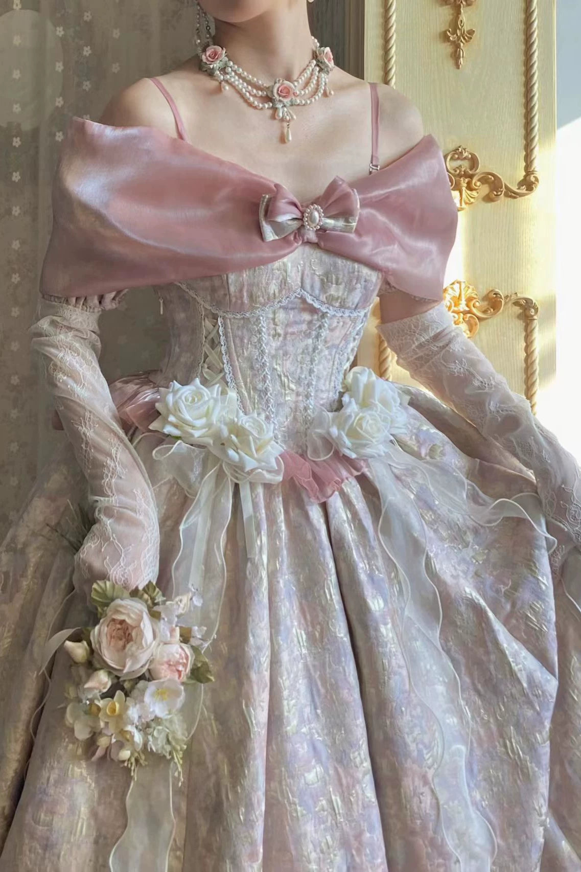 A close-up of a person wearing an ornate vintage-style ball gown with a pale pink satin cape-like shoulder drape, white floral appliqués, sheer long gloves, and layered pearl necklaces in an indoor setting with warm gold-toned wall decor.