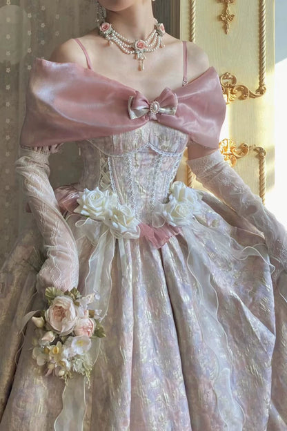 A close-up of a person wearing an ornate vintage-style ball gown with a pale pink satin cape-like shoulder drape, white floral appliqués, sheer long gloves, and layered pearl necklaces in an indoor setting with warm gold-toned wall decor.