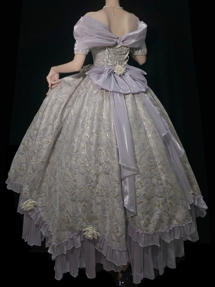 A voluminous off-the-shoulder ball gown with a lavender satin bow and a heavily embroidered beige lace overlay, shown from the back against a black background.