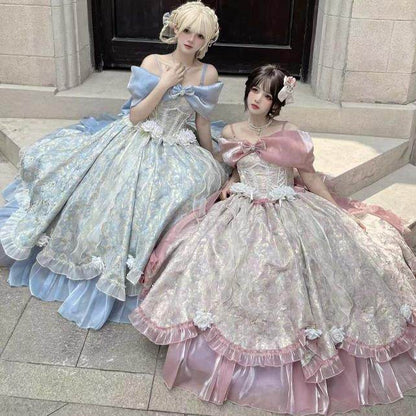 Two women posing in elaborate, voluminous Lolita-style ballgowns. The woman on the left wears a light blue gown with a pale blue satin off-shoulder strap and a cream corset bodice.