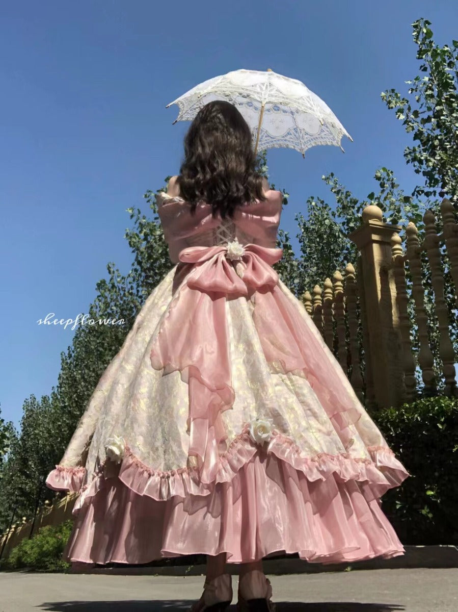 A person viewed from behind wearing an elaborate pink and cream Lolita-style dress with a large bow at the back, holding a white lace parasol outdoors under a blue sky.