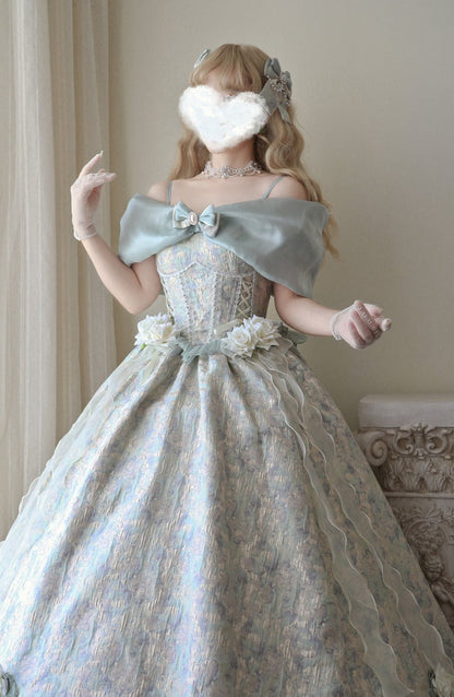 A woman wearing an ornate off-the-shoulder ball gown with a large bow, floral waist detail, and voluminous lace skirt, posed indoors against a light curtain and wall.