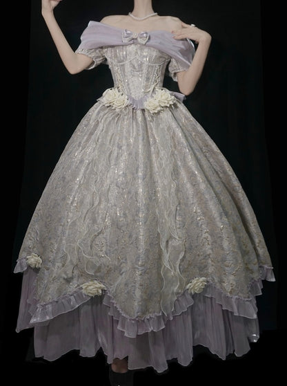 A voluminous off-the-shoulder ball gown with a heavily embroidered bodice, floral appliqués at the waist and hem, and layered ruffled tulle in a pale lavender and champagne tone, shown against a black background.