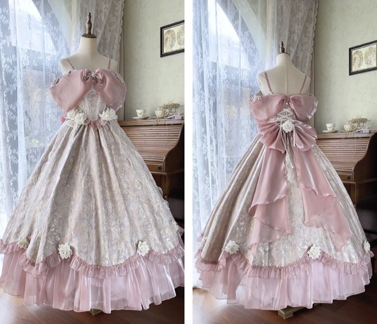 A long formal dress displayed on a mannequin in a room with lace curtains and a wooden cabinet. The dress has a pale pink and white floral pattern, off-the-shoulder straps, a large pink bow at the waist, and a layered ruffled hem.