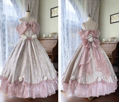 A long formal dress displayed on a mannequin in a room with lace curtains and a wooden cabinet. The dress has a pale pink and white floral pattern, off-the-shoulder straps, a large pink bow at the waist, and a layered ruffled hem.