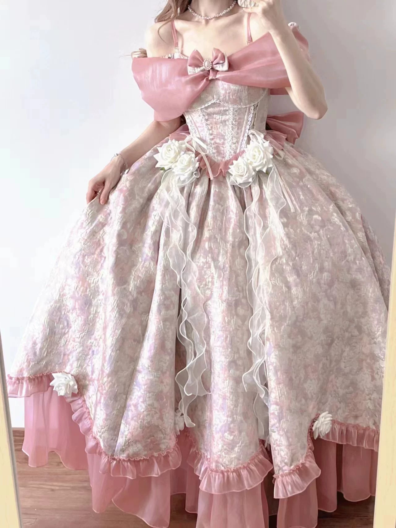 A pink and white floral Lolita-style ball gown with a large off-the-shoulder bow, corseted bodice, and voluminous ruffled skirt, shown on a model in front of a plain indoor wall.