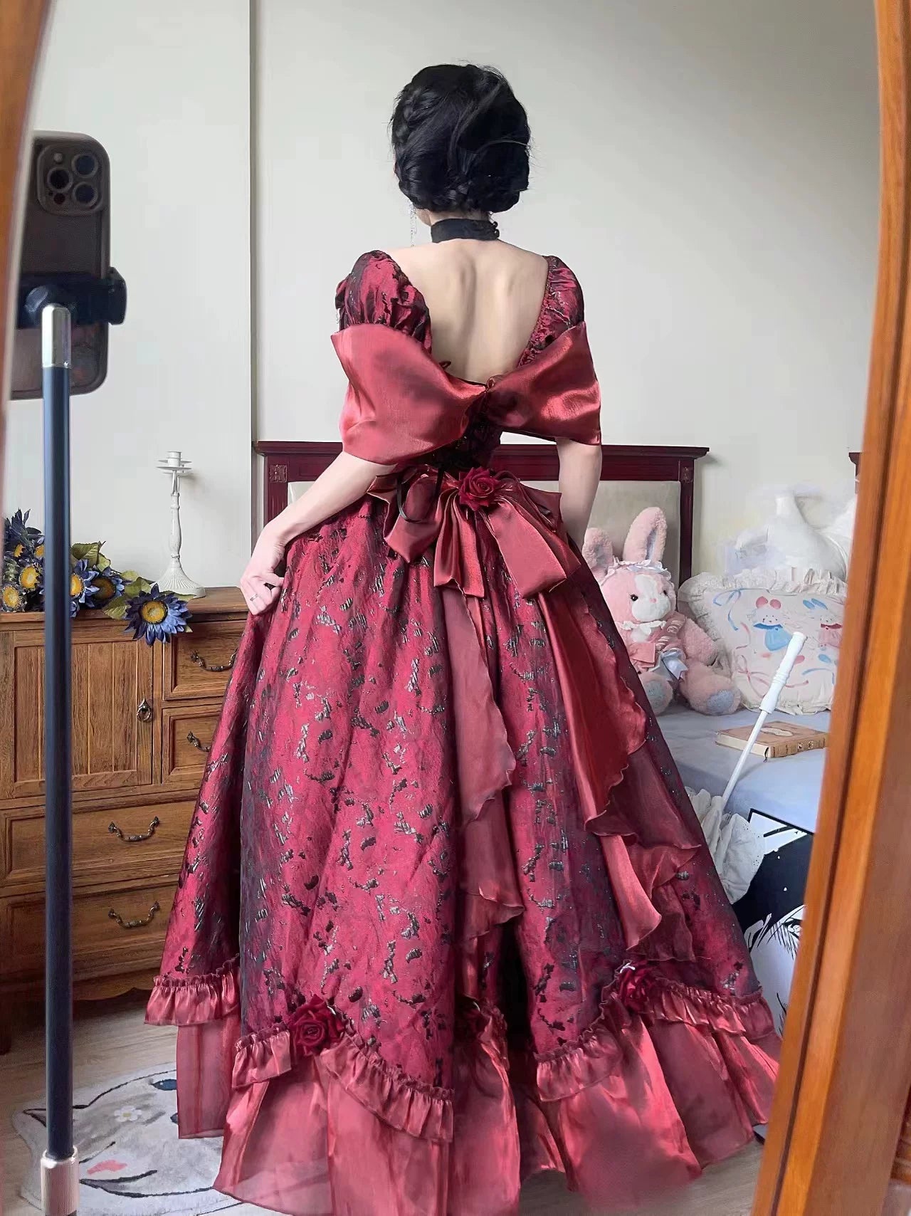 A person viewed from behind wearing an ornate floor-length ball gown with a large open back, voluminous ruffled skirt, and oversized bow details. The dress is a deep wine red with subtle floral embroidery and is photographed indoors in front of a mirror, with a phone stand visible on the left.