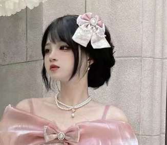 A woman with short black hair models a pink and white bow hair accessory, wearing a shiny pink top with a matching bow detail. She is posed against a light gray stone wall.